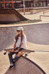 Beautiful young woman in sunglacces and cap is sitting at skatepark on the ramp with her longboard. 