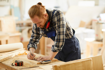 Portrait of modern carpenter marking wood while working in joinery lit by sunlight, copy space