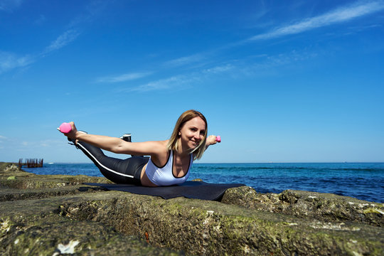 Horizontal photo of a beautiful woman who does sports exercises on the beach of the ocean or sea. The girl is lying on her stomach with widely spread arms and trains her back.