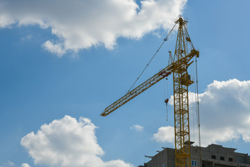 Fototapeta na wymiar high-rise construction crane with a long arrow of yellow color against the blue sky over a new multi-storey building of concrete and brick under construction