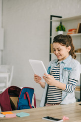 adorable smiling kid holding digital tablet while standing near desk at home