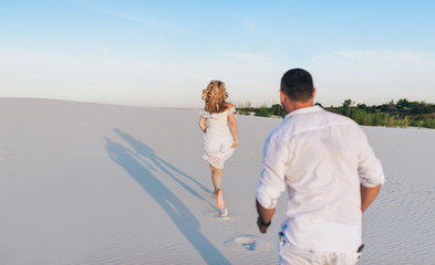 A young man runs after a beautiful blonde girl along the sand of a beach. White shirt, dress and shorts. Concept for love, vacation and honeymoon. Sunset.