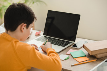 schoolboy doing homework while sitting at desk near laptop with blank screen