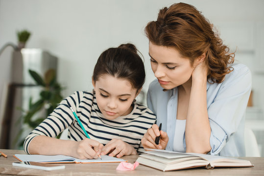 attractive woman helping concentrated daughter doing schoolwork at home
