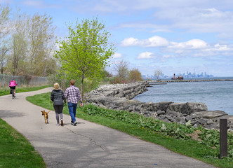 A couple walks a dog as a cyclist rides by on Toronto's waterfront trail route beside Lake Ontario - obrazy, fototapety, plakaty