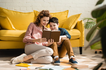 smiling mother and son sitting on floor near yellow sofa and reading book together