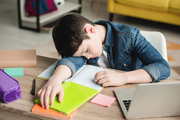adorable exhausted boy sleeping at table near copy books and laptop
