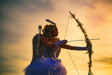 Silhouette of a cupid. Side view of teen girl archer against sunset. Little Cupid girl aiming at...