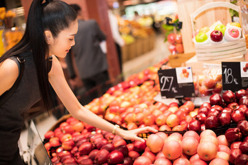 Black long hair adult Asian Woman in gray dress select pick up red cherry from bulk cherry box rack from supermarket, selective focus. Concept decision making best choice from many good things