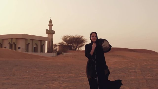 Portrait of a young Arab woman wearing traditional black clothing during beautiful sunset over the desert. Mosque on the background