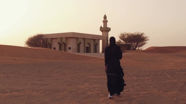 Portrait of a young Arab woman wearing traditional black clothing during beautiful sunset over the desert. Mosque on the background