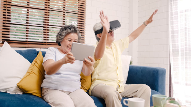 Asian elderly couple using tablet and virtual reality simulator playing games in living room, couple feeling happy using time together lying on sofa at home. Lifestyle Senior family at home concept.