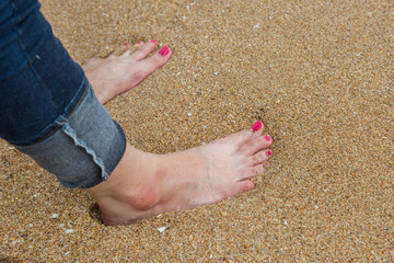 Female feet with red pedicure wear blue jean stand on the beach sand