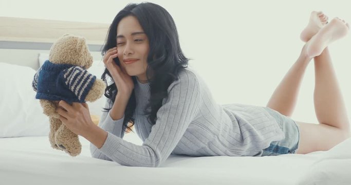 Beautiful Asian woman lie on her stomach on bed and playing with teddy bear by happy mood. 