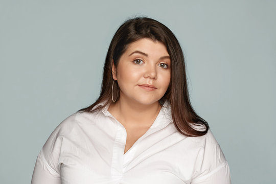 Horizontal Studio Portrait Of Adorable Beautiful Young Overweight European Woman With Loose Black Hair And Chubby Cheeks Wearing Round Earrings And Xxl White Shirt Posing At Blank Wall, Smiling