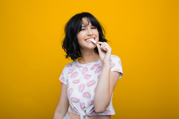 Fototapeta premium Lovely sexy young female looking at camera smiling while biting from a chewing gum against a yellow background.