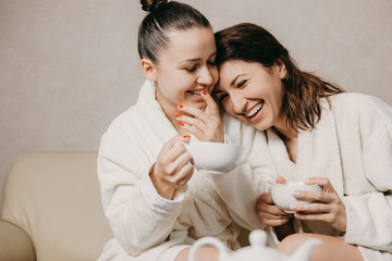 Obraz na płótnie Canvas Two beautiful girlfriends having fun dressed in bathrobes after spa procedures laughing while one is leaning her head her girlfriend.