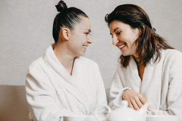 Side view portrait of two lovely young caucasian woman having fun laughing sitting in a wellness spa massage dressed in bathrobes after procedures.