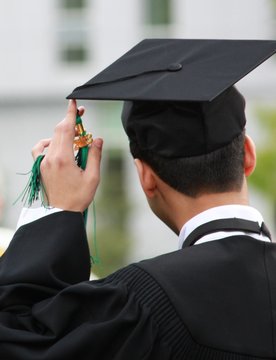 Graduate image from backside, wearing a graduation cap and gown. Left hand touching the tassel