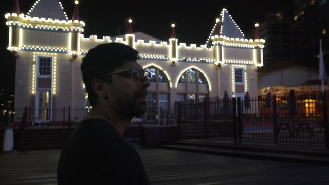 Man Walking on Wharf Near Theme Park at Night, Bright Fairy Lights in Background