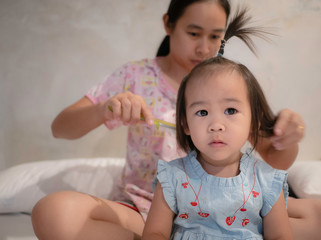 Charming Asian little girl is looking at camera and smiling while her young mother is combing daughter's hair at morning in the room.