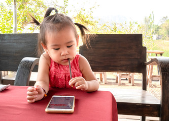 Asian little child girl sitting on wooden chair while looking smartphone in the park, waiting her mom.