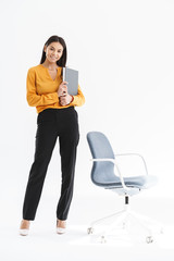 Full length portrait of charming young businesswoman dressed in elegant wear holding paper folder and standing by chair in office