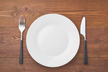 clean empty white plate, fork and knife on brown wooden table, copy space, mock up, top view.