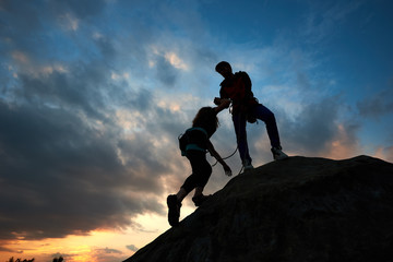 Silhouettes of couple man and woman climbers helping each other hike up mountain at dramatic sunrise background. Business, teamwork, success, goal, education, couple, love and help concept. Dark view. - obrazy, fototapety, plakaty