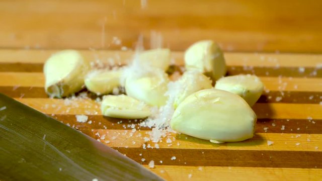 Salt is being dropped on garlic cloves on a wooden cutting board, being ready to get minced.