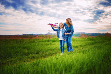 Young woman and girl in denim clothes having fun with small plane on green lawn in summer day