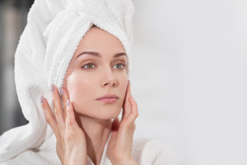 Attractive middle-aged blond woman with white towel on her head and in bathrobe standing in the bathroom by the mirror. She touches skin and smiles.