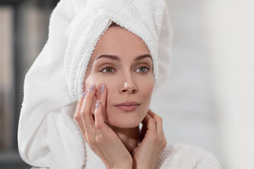 Attractive middle-aged blond woman with white towel on her head and in bathrobe standing in the bathroom by the mirror. She touches skin and smiles.