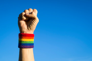 Gay protestor punching the air in defiance with gay pride rainbow colors wristband against bright blue sky