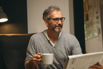 middle aged bearded man reading newspaper and having cup of coffee in his home