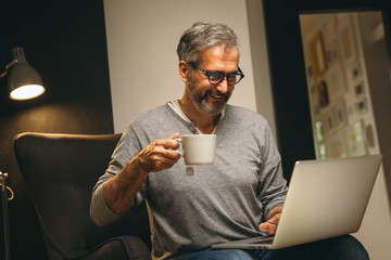 middle aged man using laptop and drinking tea relaxed in his sofa at home