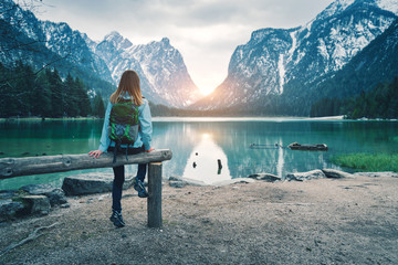 Fototapeta Young woman with backpack is sitting on the coast of mountain lake at sunset in spring. Travel in Italy. Landscape with slim girl, reflection in water, snowy rocks, green trees. Vintage toning obraz