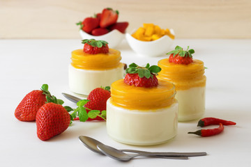Traditional Italian sweet dessert Panna cotta with mango jelly with touch of red hot chili Piri-Piri served in jars on white wooden table and embellished with fresh real strawberrries and mint.