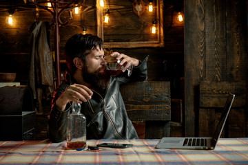 Fototapeta na wymiar Drinking man. Man with beard holds glass brandy. Man holding a glass of whisky. Sipping whiskey. Degustation, tasting. Macho is drinking whiskey during working on his laptop.