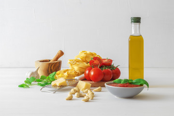 Table with ingredients to make tomato pesto. Tomatoes, garlic, fresh oregano and basil herbs, bottle of olive oil, few cashew nuts, parmesan cheese, cheese grater, wooden mortar and homemade pasta.