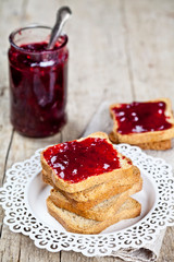 Toasted cereal bread slices on white plate and jar with homemade cherry jam closeup on rustic wooden table background.