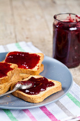 Fresh toasted cereal bread slices with homemade cherry jam in jar and spoon on ceramic plate closeup on linen napkin on rustic wooden table background.
