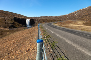 Human and nature: clean water of waterfall and asphalt road on a stony rocky mountain landscape of Iceland