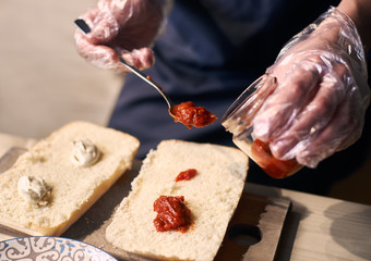 Cropped hands with spoon taking sauce from glass jar. Putting red and white sauce over large pieces of bread. Preparing sandwich by traditional Italian recipe. Close up view of dish in cooking process