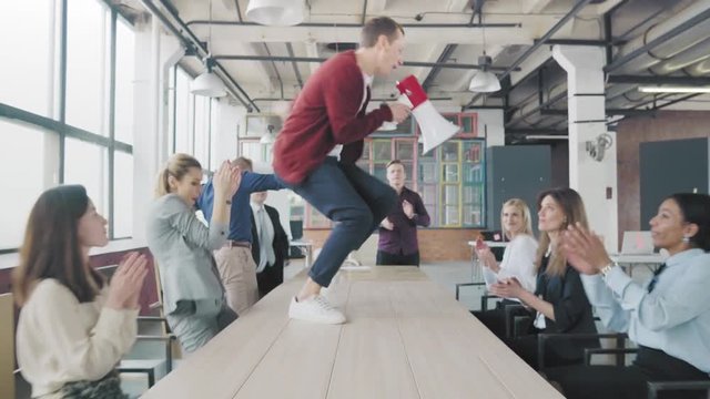 Manager start to dance on the table, sings into a megaphone, accelerates and slide on lap. Colleagues blow up flappers with confetti. Employees celebrate success. Corporate party business team