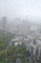 City buildings and the green of trees are just visible through a rain-smeared window. Rain drops are running down the pane.