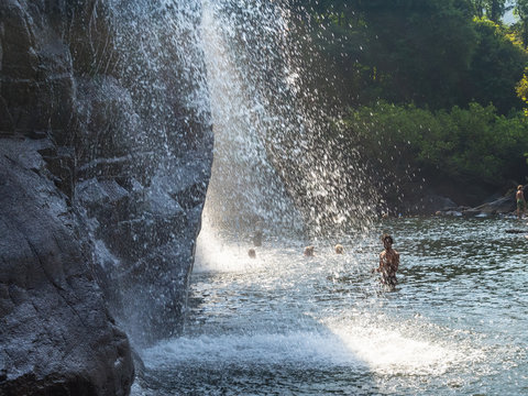 A Happy Middle-aged Man Stands Under A Waterfall In The Jungle. Streams Of Water Falling From Above And Sparkle In The Sun. A Male Baths And Enjoys His Relax.