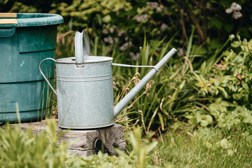Old metal watering can standing on an weathered wooden bar in front of a water container in a green natural springtime garden
