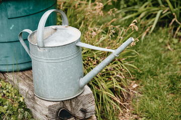 Old metal watering can standing on an weathered wooden bar in front of a water container in a green natural springtime garden