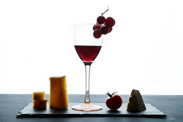 A few pieces of cheese and cherry tomatoes next to a glass of red wine filled with grapes on a white background